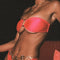 A model wearing a strapless, tie-back bandeau bikini top in magenta hue, with a vintage gold ring detail, posing in a seated position.
